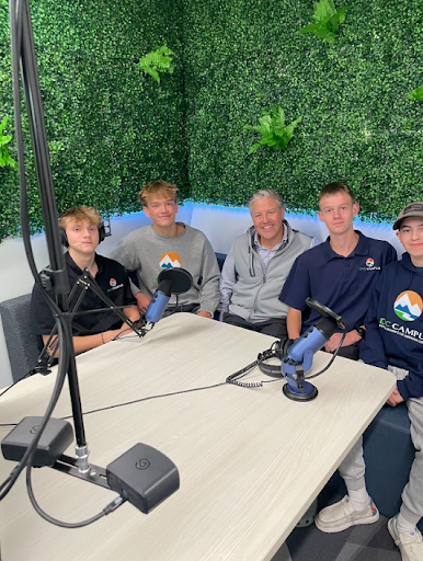 Four young men and one older man are seated around a table with microphones, posing for a photo in a podcast recording studio.