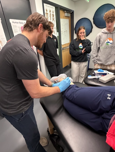 Student applies treatment to another student's ankle on a table, as a group of peers watches and observes.