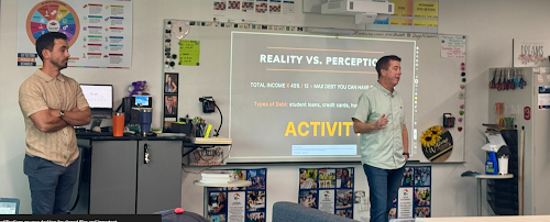 Two men stand in a classroom, one presenting a slide titled "Reality vs. Perception" on a projection screen.