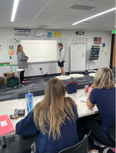 A teacher and a male student stand near a projection screen while two female students sit at a table in the foreground.