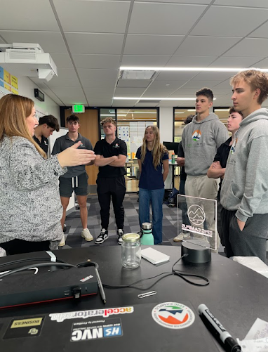 A female teacher speaks to a small group of high school students gathered around a table.