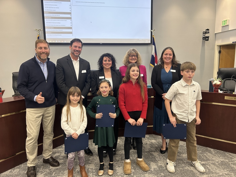 Five adults and four elementary students holding certificates pose together in a meeting room.