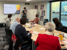 A student presents a slide show to a group of adults seated around a conference table.