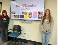 Two female students stand on either side of a screen displaying "The EPICenter" and food images.