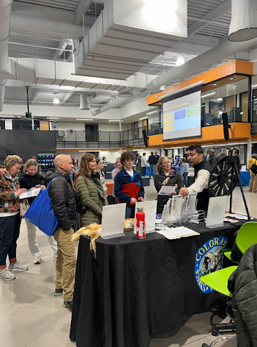 People gather around a table with a "Colorado" logo sign at an indoor event.