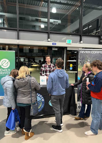 A group of people stand around a table with "River Corps" and "Emily Griffith" banners in a large room.