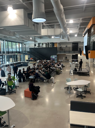An overhead shot of a large, modern indoor space with many students seated for a presentation.