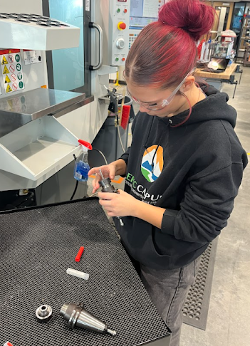 A student with bright red hair works on a metal component next to a large piece of machinery.