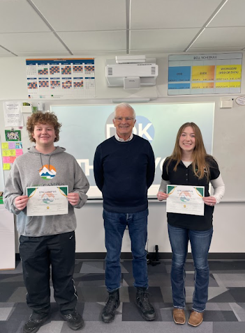An older man stands between two students holding certificates in a classroom.