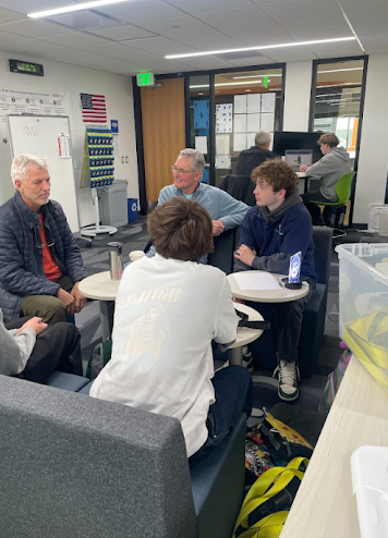 A student group and two older mentors discuss a topic around low-slung, rounded seating.