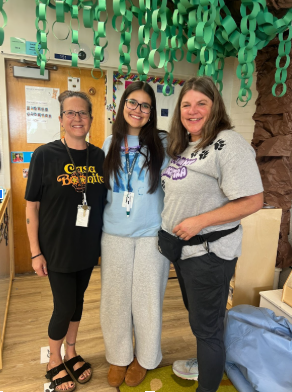 Three smiling women pose together in a classroom under hanging green paper chains and next to a paper tree decoration.