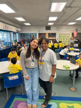 Two women smiling in a busy classroom as students in yellow shirts work at circular tables in the background.