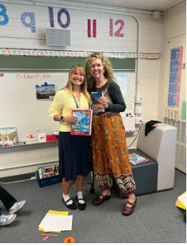 Two women smiling and holding books in a classroom in front of a whiteboard and number line.