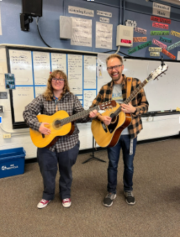 A man and a woman smiling and posing while holding acoustic guitars in a music classroom.