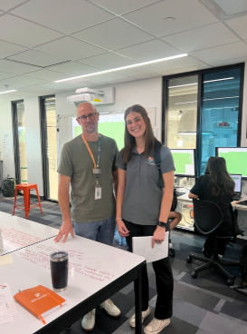 A man and woman posing in a modern computer lab with monitors and a collaborative whiteboard table.
