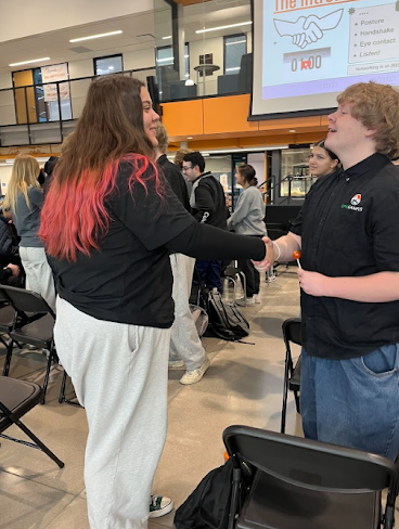 Two students practice a professional handshake in a large common area during a networking skills workshop.
