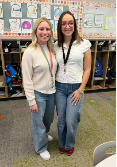 Two smiling women stand together in a classroom in front of student artwork and cubbies filled with backpacks.