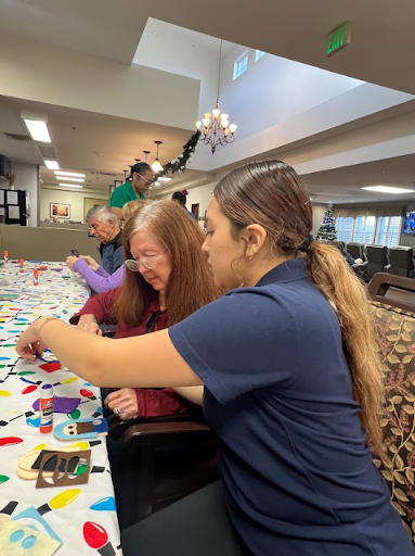 A young woman assists a senior with a colorful craft project at a long table in a bright community room.