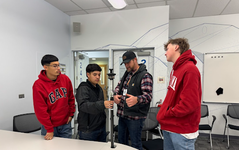 An instructor shows three students how to use a vertical surveying tool inside a classroom.