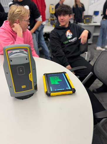 Two students sit at a table with a Trimble X9 scanner and a tablet displaying a 3D green floor plan map.