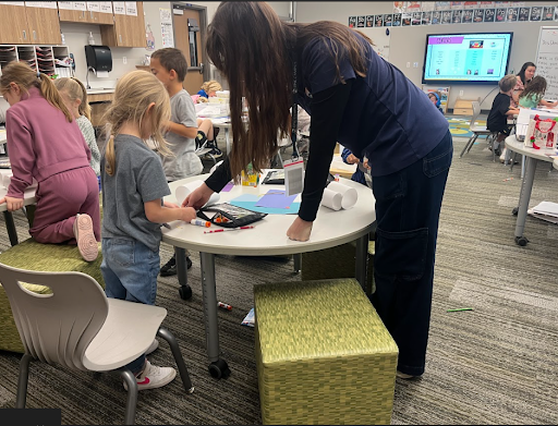 A student teacher leans over a round table to assist a young child with a paper craft project in a classroom.