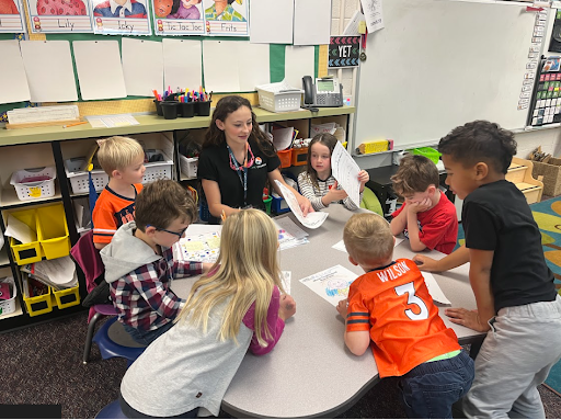 A student teacher sits at a table with six elementary children, showing them worksheets in a colorful classroom.