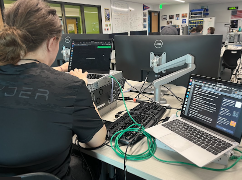 Student using a laptop to configure a server rack with green ethernet cables in a tech lab.