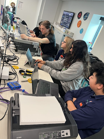 Group of students working on laptops and networking hardware in a computer science classroom.