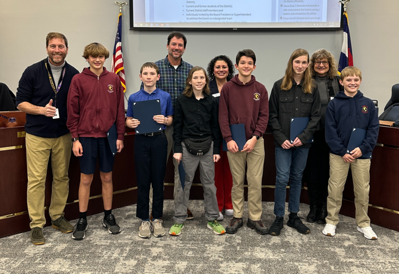 Six students hold awards and stand with four adults in front of a large screen at a Littleton Public Schools Board meeting.