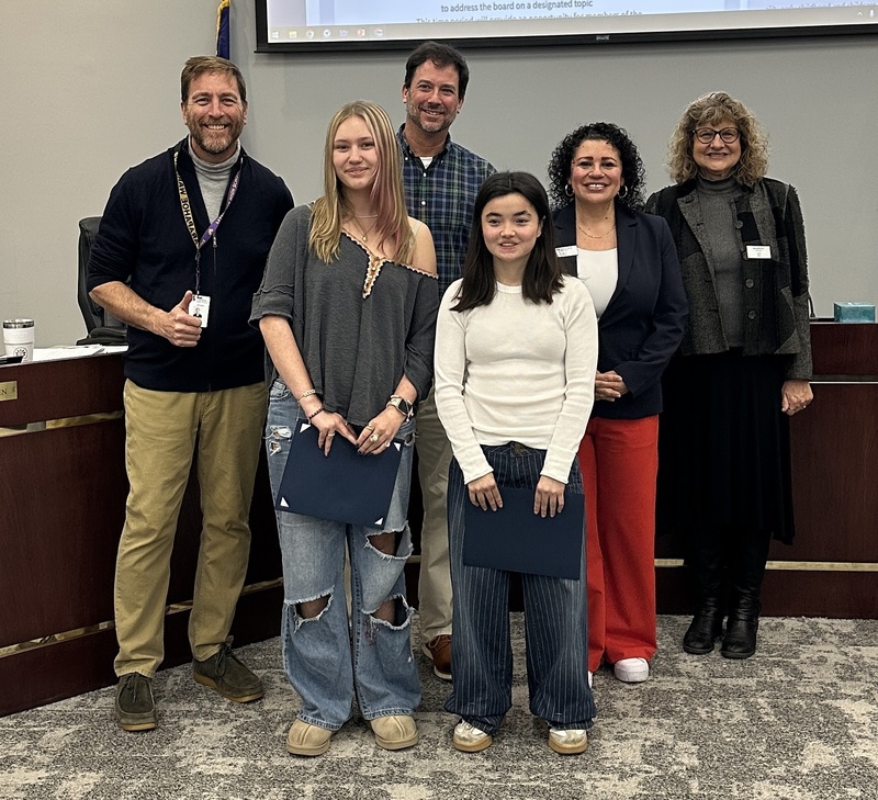 Two students hold certificates while standing with four adults in front of a presentation screen at a board meeting.