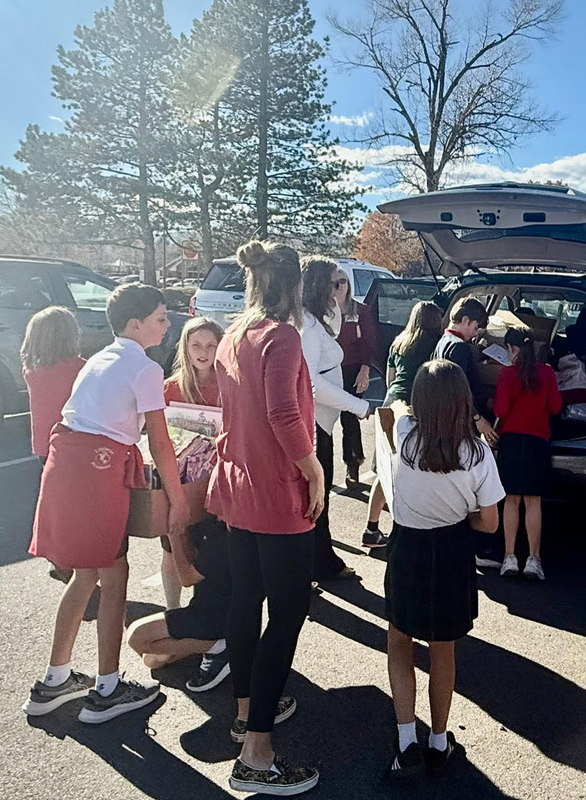 Students and adults load boxes of books into the back of a car in a sunny parking lot.