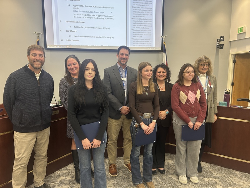 Three students holding award folders stand with LPS officials in front of a board meeting screen for Student Spotlight.
