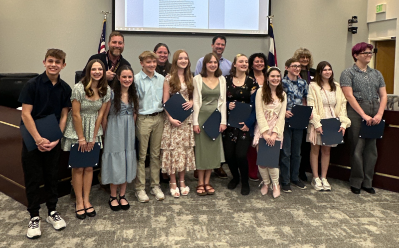 A large group of students stand together holding blue award folders at a Littleton Public Schools board meeting.
