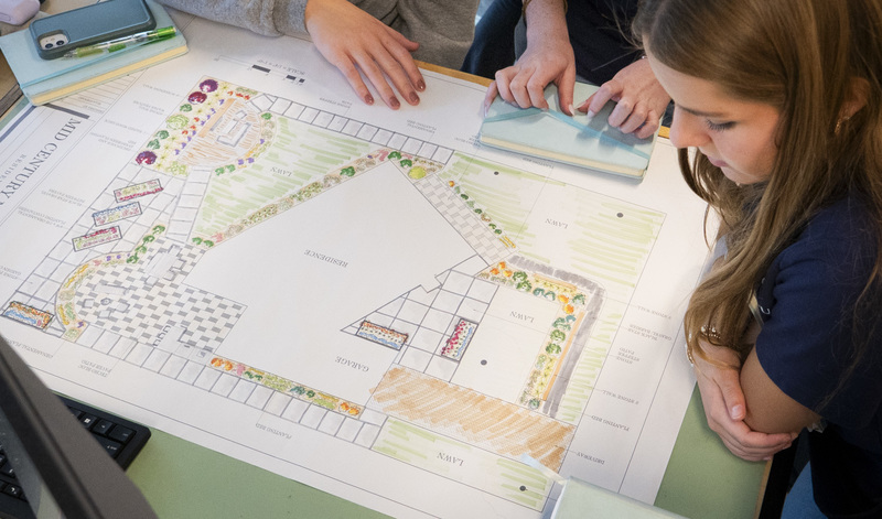 A student reviews a large architectural landscape blueprint titled "Mid Century Residence" on a desk with classmates.