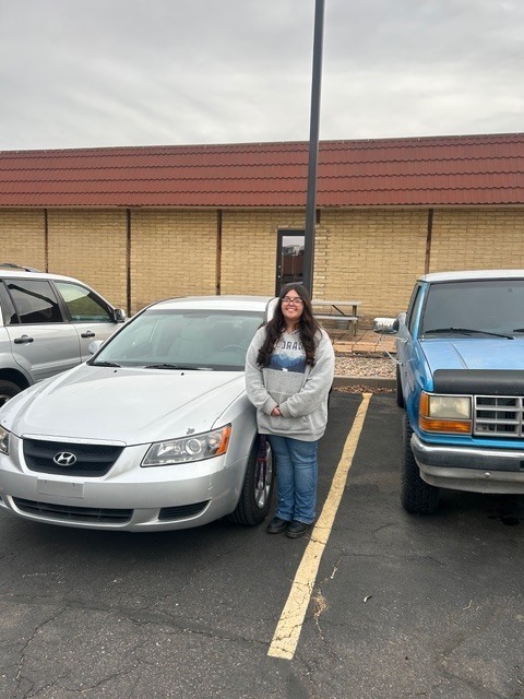 A smiling person in a grey sweatshirt stands next to a silver Hyundai in a parking lot between two other vehicles.