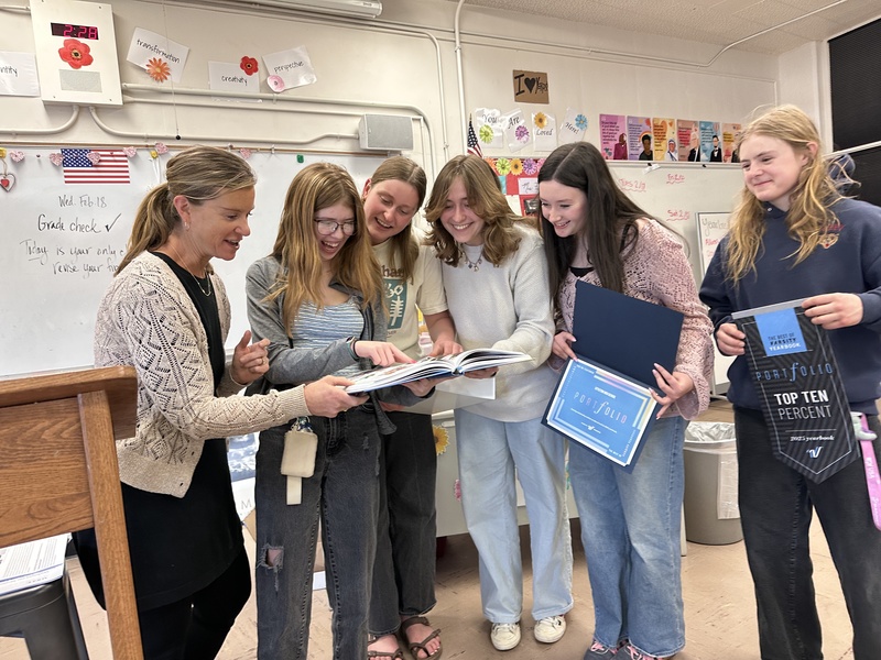 A teacher and students gather around to look at a yearbook together while holding an award certificate.