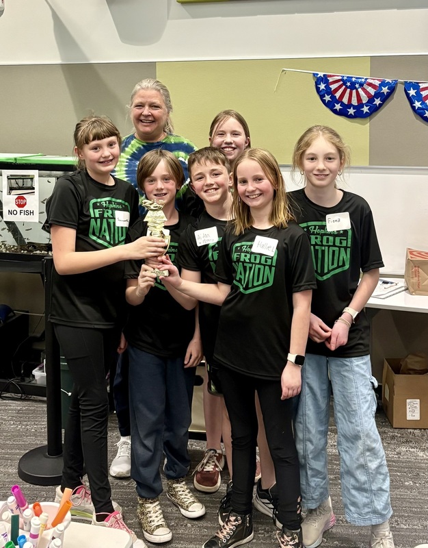 6 students and their coach from Hopkins Elementary wear matching Frog Nation tshirts and hold a Battle of the Books tropy.