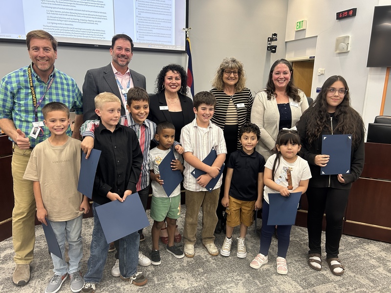 Elementary-aged students and staff smile while holding blue award folders during a Board of Education meeting.
