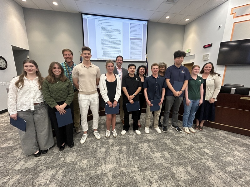 High school students stand with staff in a meeting room, holding blue award folders for a student spotlight.
