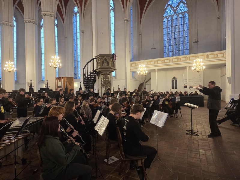 Student orchestra performs in a white Gothic cathedral with tall arched windows and a spiral staircase pulpit.