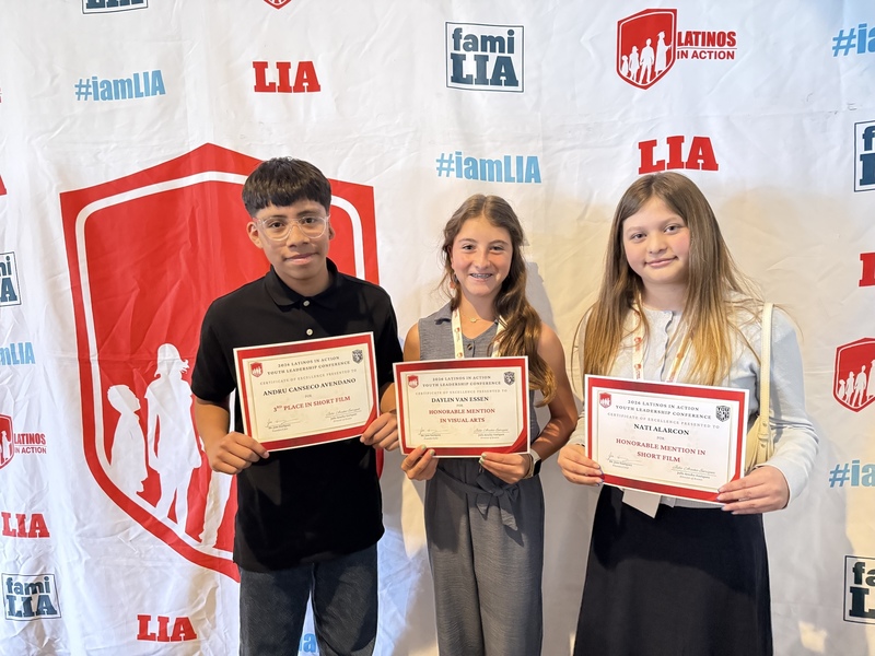 Three students smile while holding award certificates in front of a Latinos In Action backdrop.