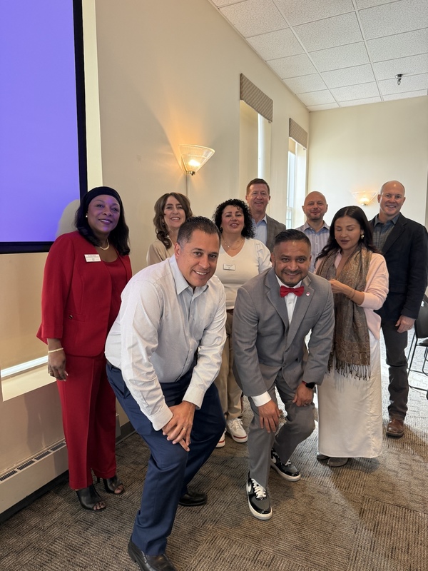 A group of diverse staff and community leaders smile for a group photo in a brightly lit meeting room.