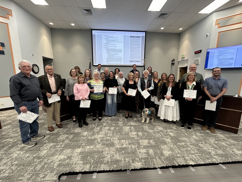 A large group of volunteers stands in a board room holding recognition certificates, accompanied by a fluffy dog.
