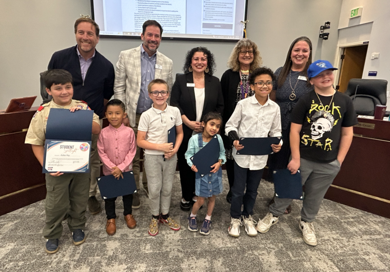 A group of young students smiles while holding award certificates with board members in a meeting room.
