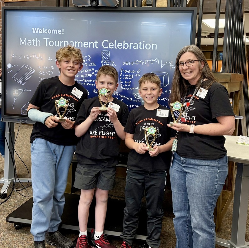 Three Ford Elementary 5th graders and their teacher smile with trophies in front of a math-themed digital display.
