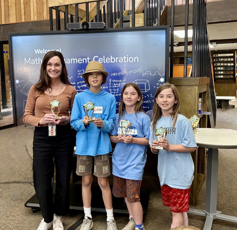 Three Little Raven 5th graders and their teacher hold trophies in front of a Math Tournament Celebration screen.