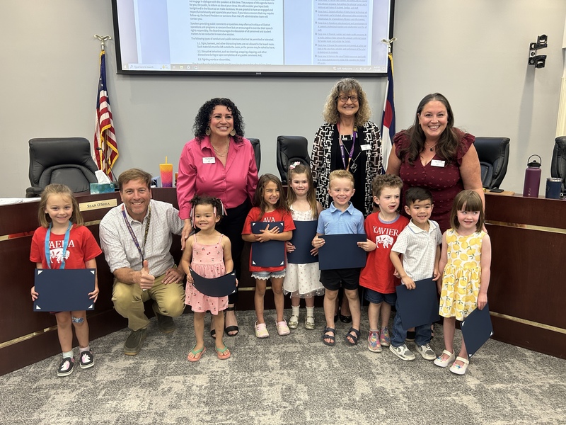 Preschool students holding blue folders pose for a photo with four LPS Board members in a meeting room.