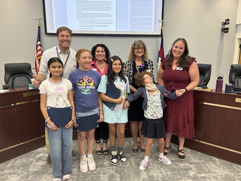 Four elementary school students stand with blue folders alongside smiling members of the LPS Board of Education in the Boardroom.