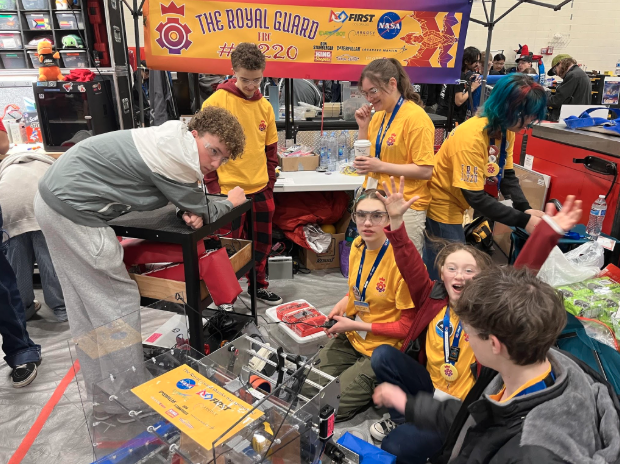AHS Robotics team members smile and work on their robot in the pit area under a banner reading The Royal Guard FRC 11220.