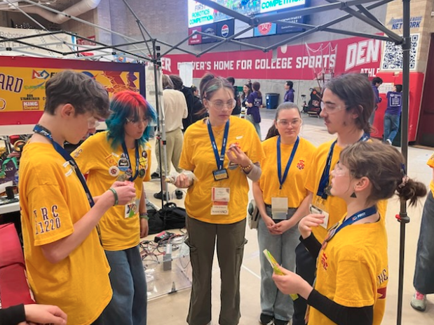 Six high school students wearing matching yellow robotics shirts and safety glasses huddle and talk at a competition.